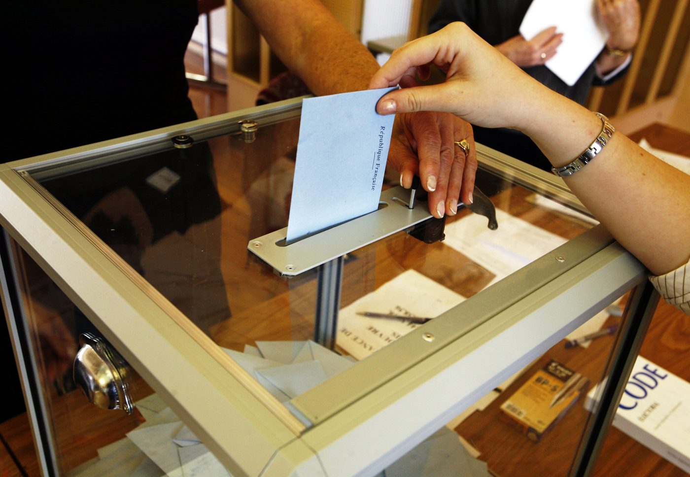 Une enveloppe de vote République française glissée dans une urne transparente, un Code électoral posé à côté